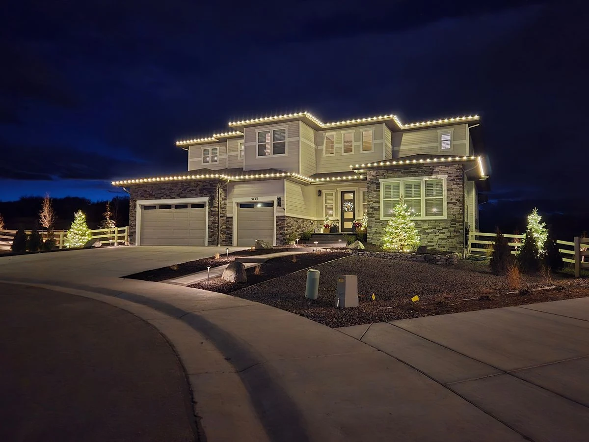 Two-story home with warm white lights and lit evergreen trees at twilight