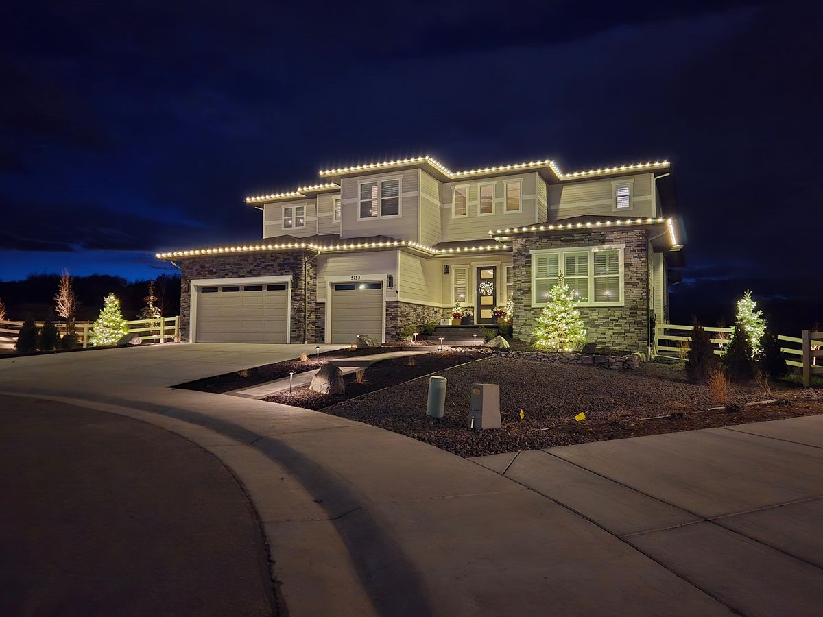 Two-story home with warm white lights and lit evergreen trees at twilight